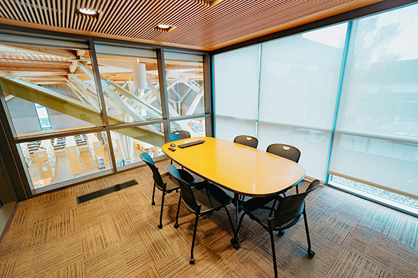 A small meeting room with a wooden oval table, seven black chairs, large windows with blinds, and carpeted floor.