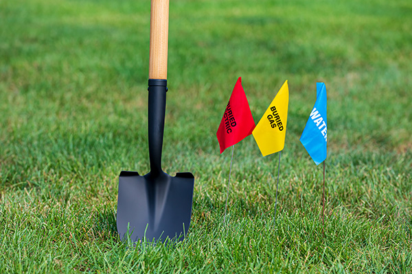 Shovel placed in grass next to three utility flags: red for electric, yellow for buried gas, and blue for water.