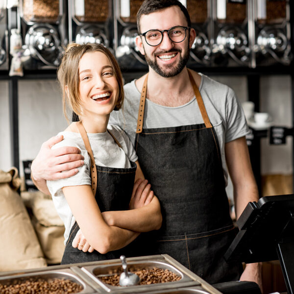 Two smiling people wearing aprons stand together in a coffee shop, surrounded by coffee beans and dispensers.