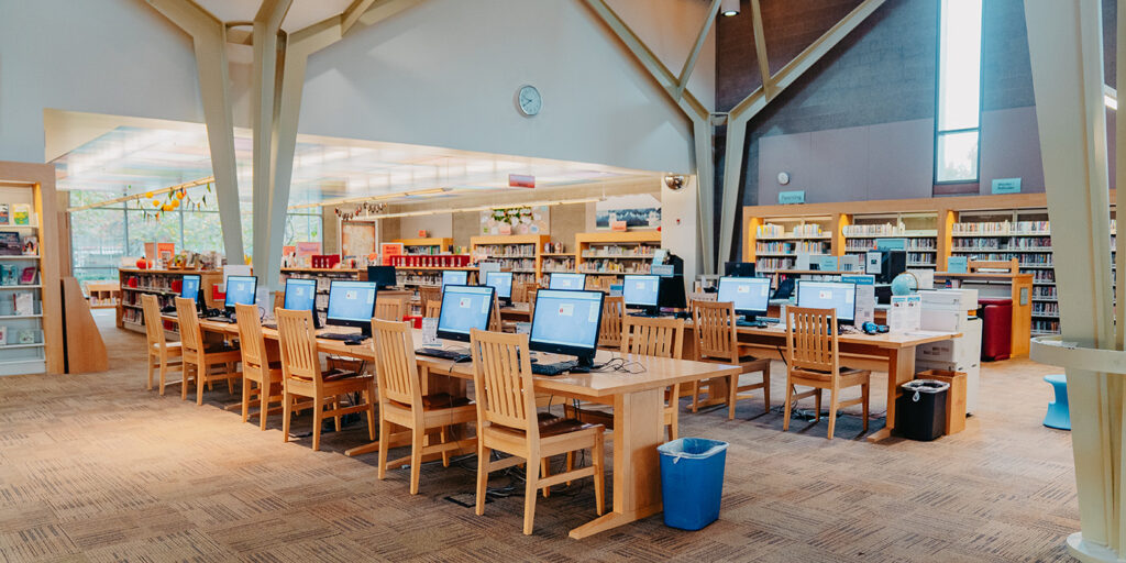 A library interior with rows of wooden chairs and computer desks, surrounded by bookshelves filled with books. A clock is visible on the wall above the computers.