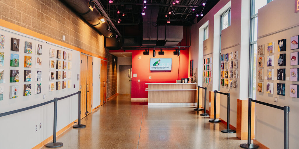 Art gallery interior with colorful artworks displayed on walls, a wooden reception desk at the end, and overhead lighting.