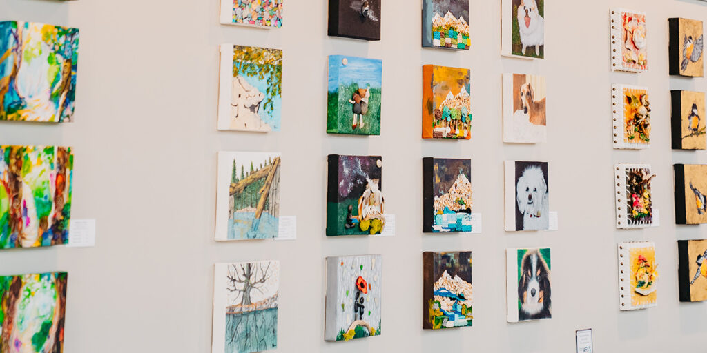 A wall displaying various small, colorful paintings featuring animals and landscapes in a gallery setting.