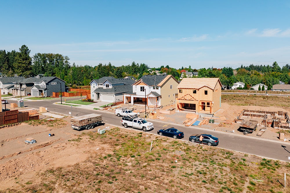 Residential construction site with several houses, two under construction, surrounded by vehicles, dirt, and building materials. Adjacent houses and trees in the background under a clear sky.