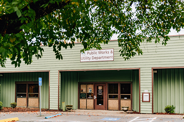 Public Works & Utility Department building with a green exterior, parking spaces in front, and a tree partially visible in the foreground.