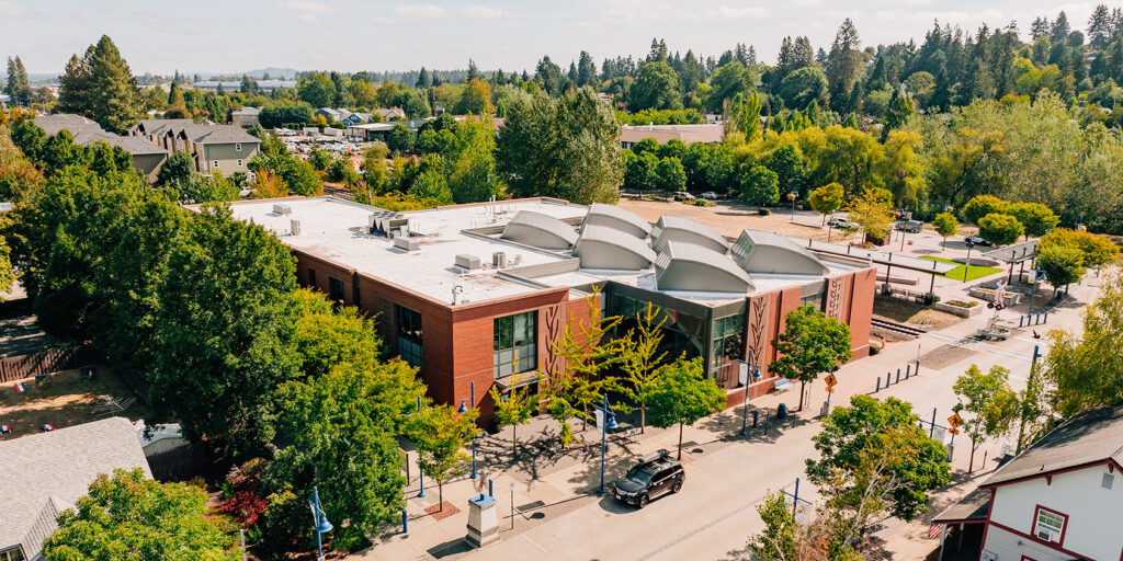 Aerial view of a brick building with a white roof surrounded by trees, situated in a suburban area with roads and parked cars nearby.