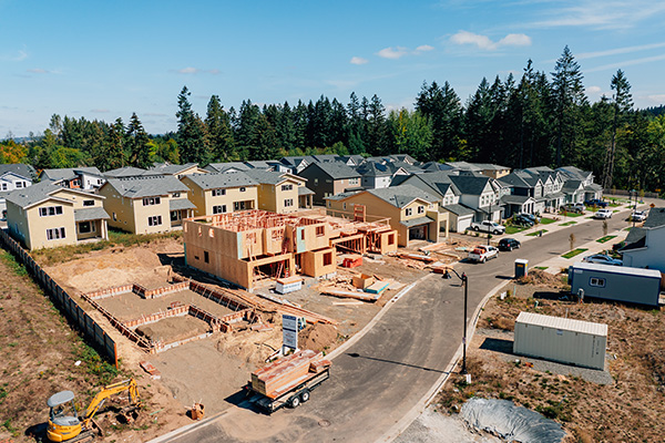 Aerial view of a suburban neighborhood with houses under construction alongside completed homes, surrounded by trees and clear skies.