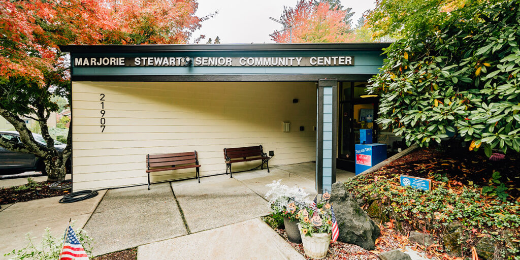 Entrance of Marjorie Stewart Senior Community Center with benches, colorful autumn trees, and an American flag decoration near the walkway.