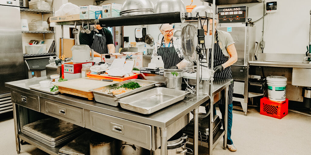 A busy commercial kitchen with two people in aprons preparing food at a stainless steel workstation, surrounded by various cooking utensils and appliances.