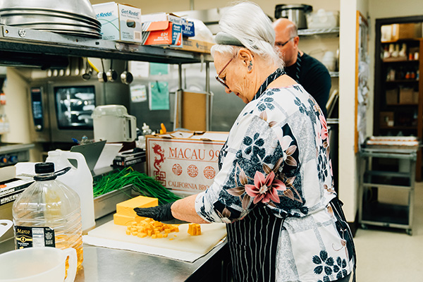 Elderly woman in a floral blouse chops cheese in a commercial kitchen, with a man in the background.