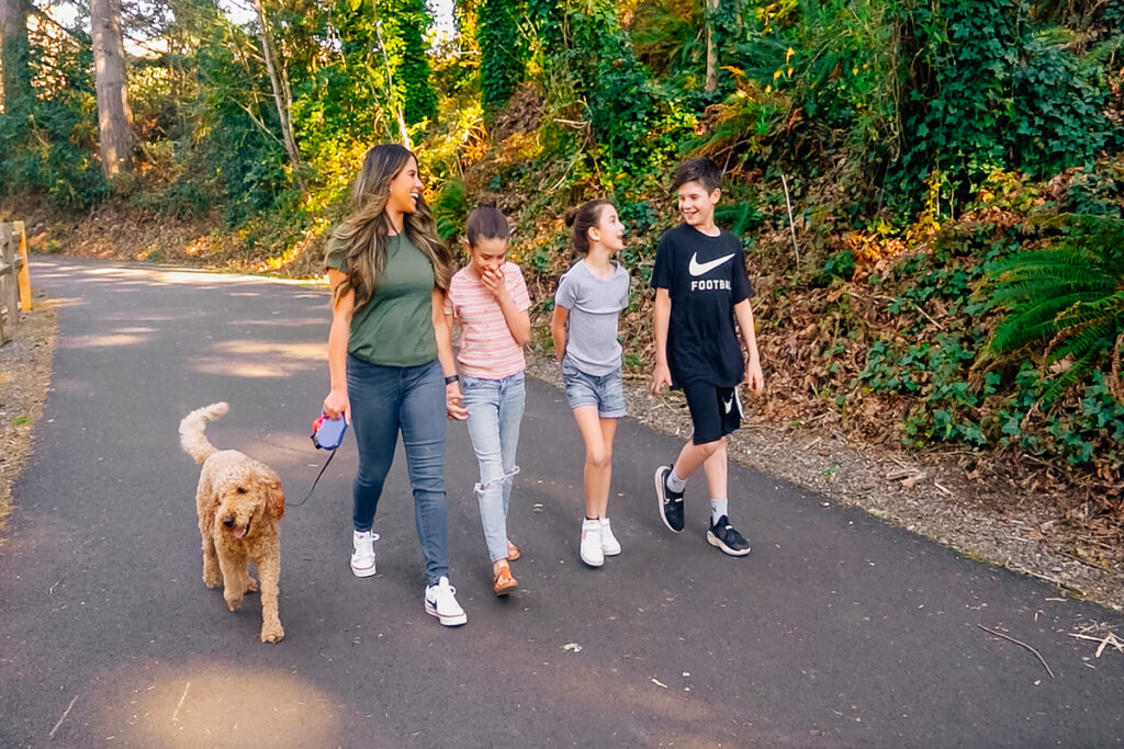 A woman and three children walk alongside a dog on a leash on a shaded path surrounded by greenery.