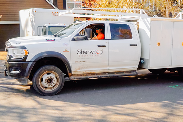 A white Sherwood Broadband utility truck with a person in an orange vest driving, parked on a residential street.