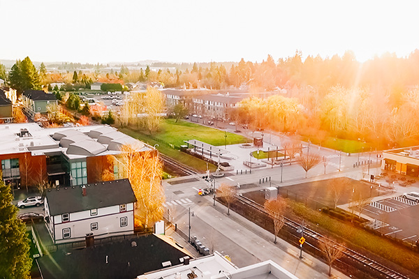 Aerial view of a small town with buildings, streets, and train tracks. The sun is setting, casting a golden light over the scene. Trees and parked cars are visible along the roads.