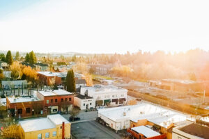 Aerial view of a small town with several buildings, streets, and trees. The sky is clear with bright sunlight casting a warm glow over the scene.