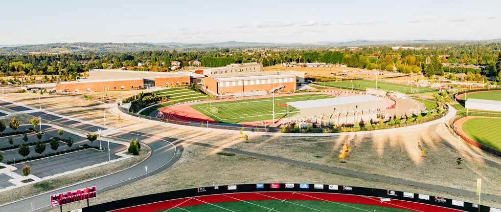 Aerial view of a high school campus with sports fields, including a track, football field, and adjacent buildings, surrounded by trees and roads under a clear sky.