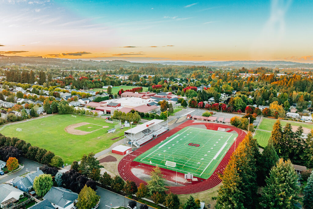 Aerial view of a high school campus with a football field, track, baseball field, and surrounding residential area under a clear sky.