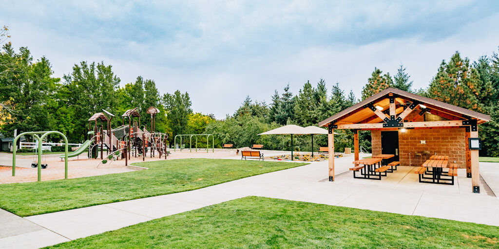 Playground with climbing structures and swings, surrounded by grass and trees. There is a covered picnic area with tables and benches nearby. Cloudy sky overhead.