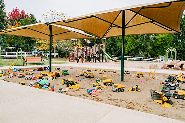 A playground sandbox filled with various toy trucks and vehicles. A large shade canopy is overhead, and a playground structure is visible in the background.