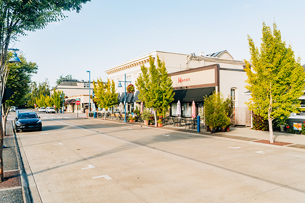 A quiet street with trees, parked cars, and buildings featuring storefronts and cafes.