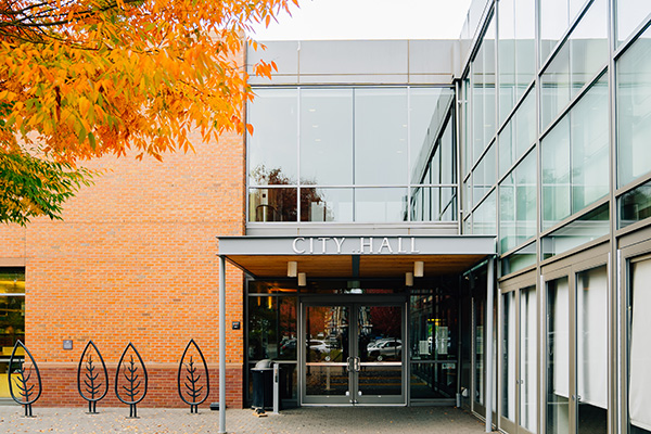 Entrance of a modern city hall building with glass windows, brick wall, and autumn trees nearby.