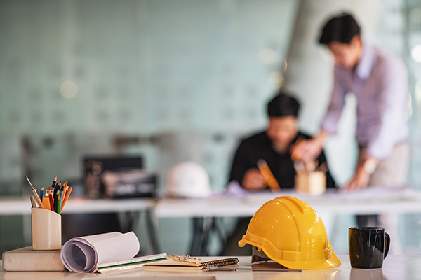 Yellow hard hat on a desk with blueprints and pencils; two people in blurred background reviewing documents.