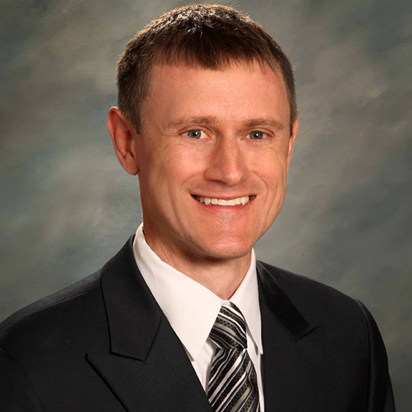 A person in a suit and tie smiles against a gray backdrop.