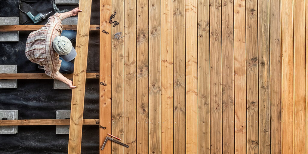 Person in a cap and plaid shirt constructing a wooden deck, holding a plank in place among beams, with tools scattered nearby.