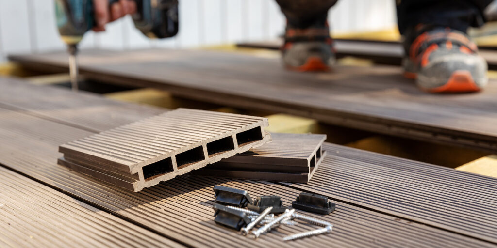 Two composite decking boards, screws, and fasteners are on a wooden deck. A person with an electric drill works in the background.