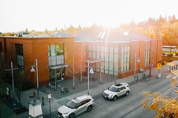 Brick building with large windows next to a street with parked cars and streetlights. Trees and a bright sky in the background.