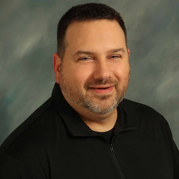 A man with short dark hair and a beard is smiling, wearing a black shirt, against a gray-blue background.
