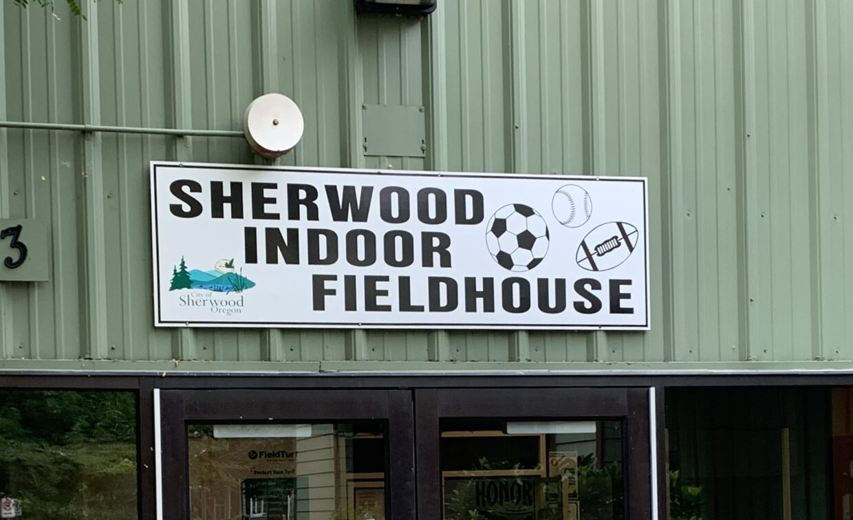 A sign reading "Sherwood Indoor Fieldhouse" with images of sports balls is mounted above glass doors on a green metal building.