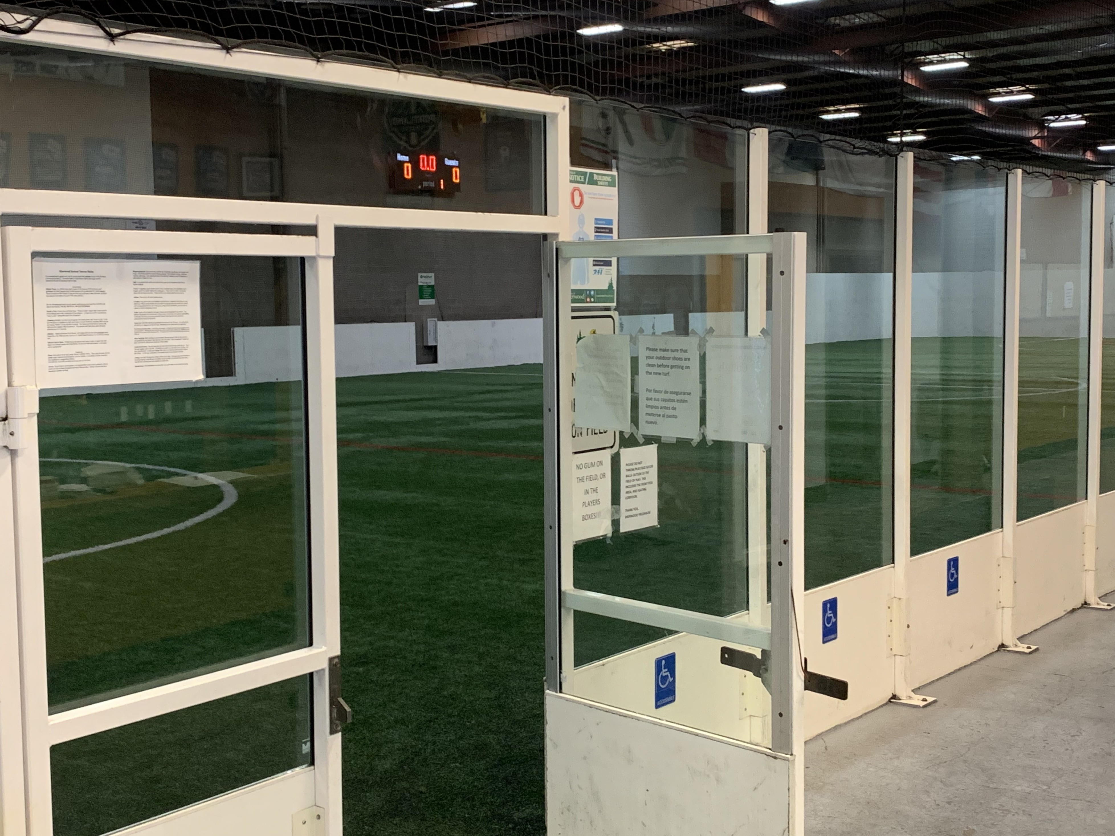 Indoor soccer field viewed through open glass doors, with accessibility signs visible and scoreboard in the background showing 0-0.