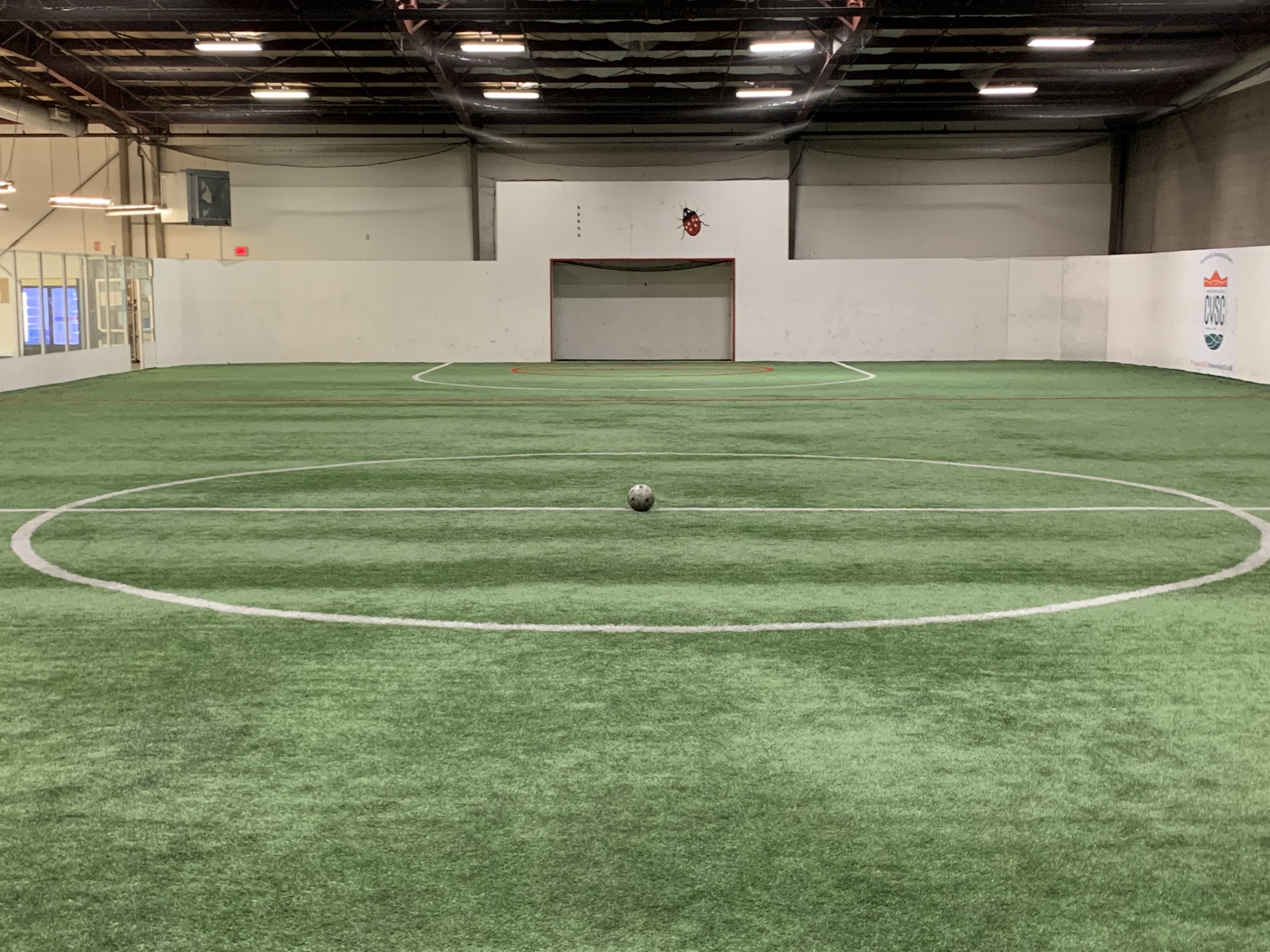 Indoor soccer field with artificial turf, centered white circle, and a soccer ball placed at midfield; goal and walls visible in the background.