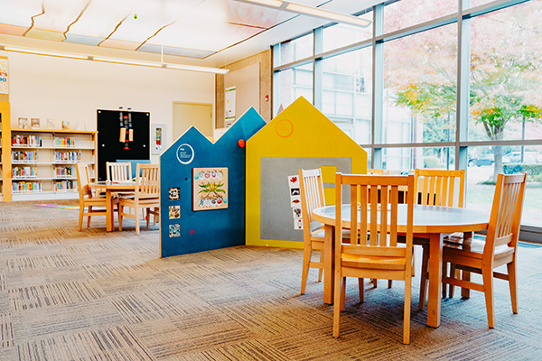 Children's reading area with small round tables, wooden chairs, colorful house-shaped panels, and large windows. Shelves with books line the background.