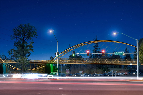 Night view of a lit bicycle bridge with a glowing arch over a busy road intersection, featuring traffic lights and streaks of car lights.