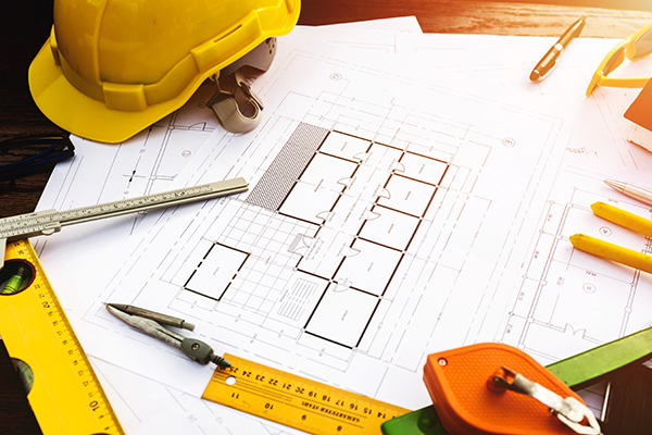 Architectural blueprints surrounded by a yellow hard hat, measuring tools, pencils, and a compass on a wooden table.