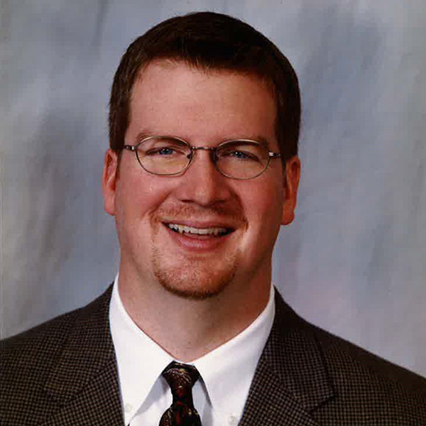 A man wearing glasses, a checkered suit, and a patterned tie is smiling against a neutral background.