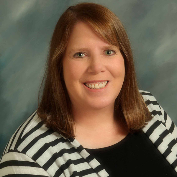 Smiling woman with shoulder-length hair wearing a black top and a black-and-white striped cardigan, against a blurred gray and blue background.