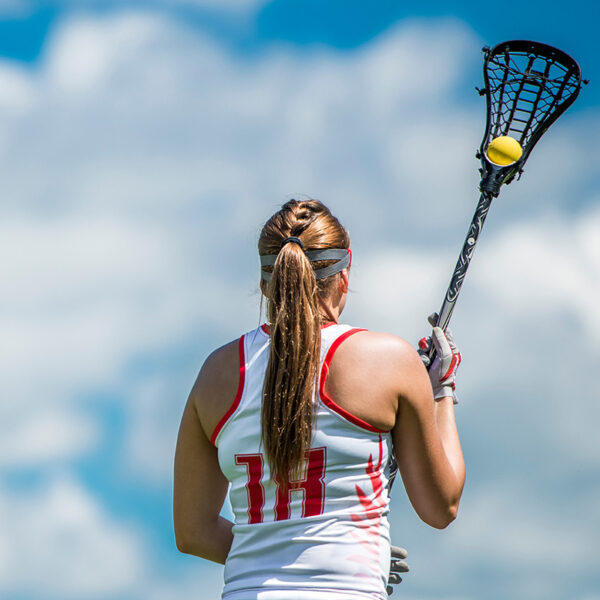 A female lacrosse player wearing a white jersey with number 11 prepares to catch a ball. She is outdoors under a cloudy blue sky.