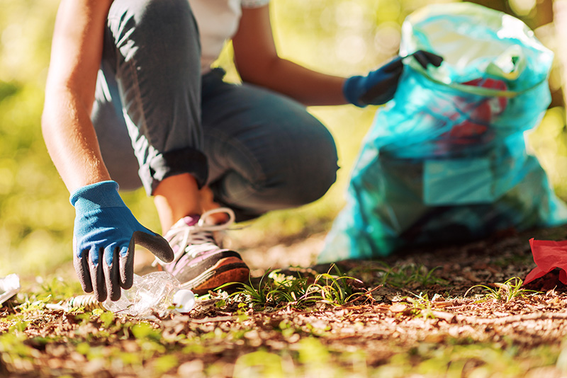 Person wearing gloves picks up litter, placing it in a blue garbage bag, outdoors on a sunny day.