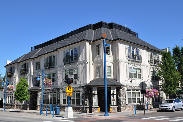 Three-story building with a sloped roof on a sunny corner, featuring large windows, hanging flower baskets, and a pedestrian crossing sign nearby.