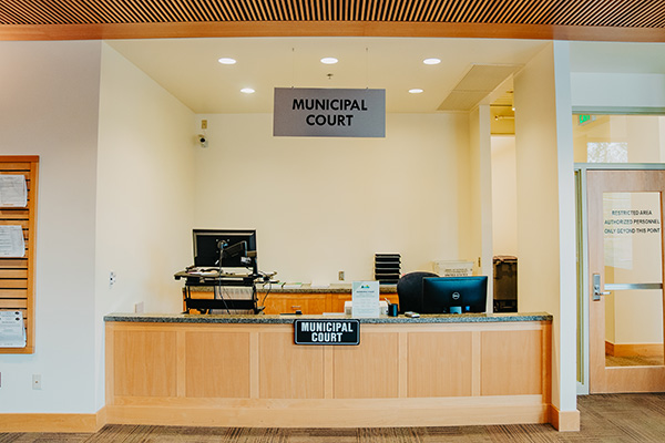 Front desk of a municipal court with a computer, chair, and signage. Wood paneling and a small stack of papers are visible.