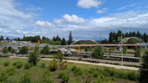 A partially constructed pedestrian bridge with white arches spans over a road, surrounded by trees, grassy areas, and construction vehicles under a partly cloudy sky.