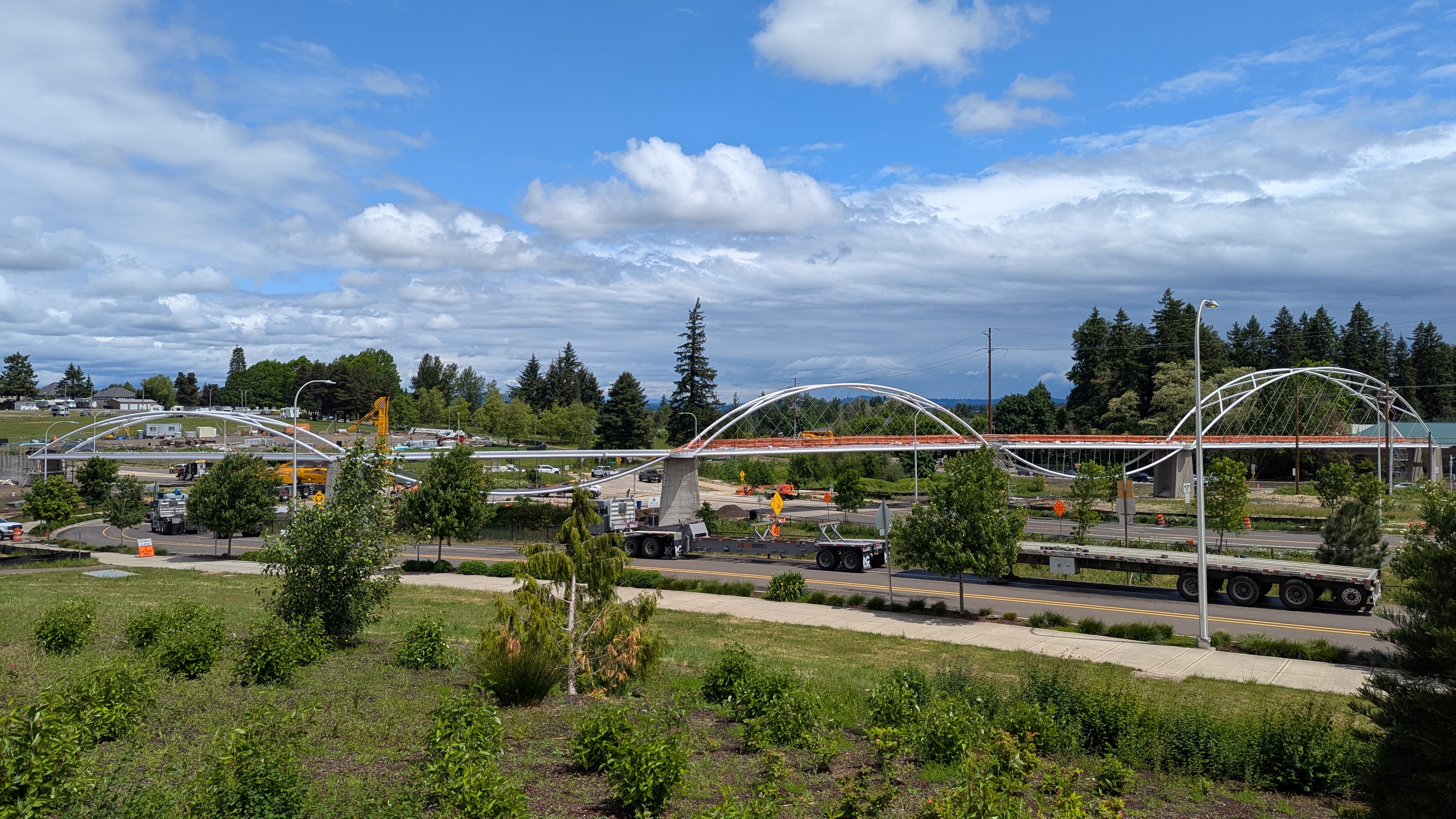 A partially constructed pedestrian bridge with white arches spans over a road, surrounded by trees, grassy areas, and construction vehicles under a partly cloudy sky.