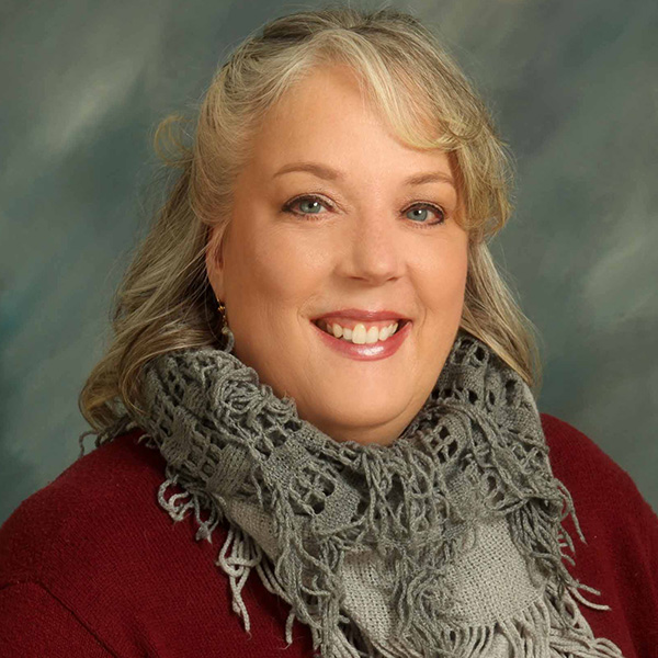 Woman with gray hair smiles, wearing a red top and grey scarf, against a blue-green background.