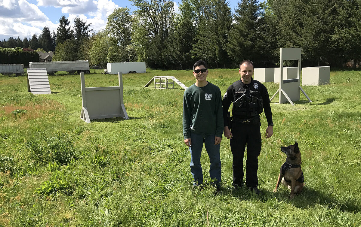 Two people stand on a grassy field, one in casual clothing and the other in a uniform. A dog is seated beside them. Training obstacles are visible in the background.