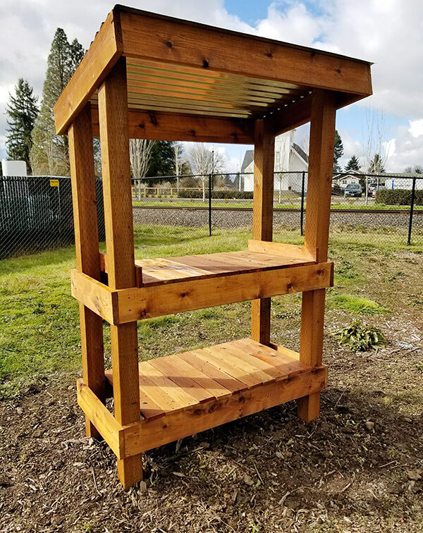 A wooden garden workstation with a roof and two shelves stands on soil in an outdoor setting with trees and a fence in the background.