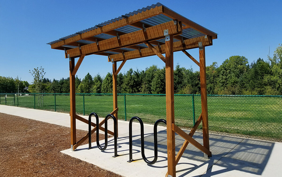 A wooden bike shelter with a corrugated metal roof over a bike rack is positioned near a fenced grassy area under a blue sky.