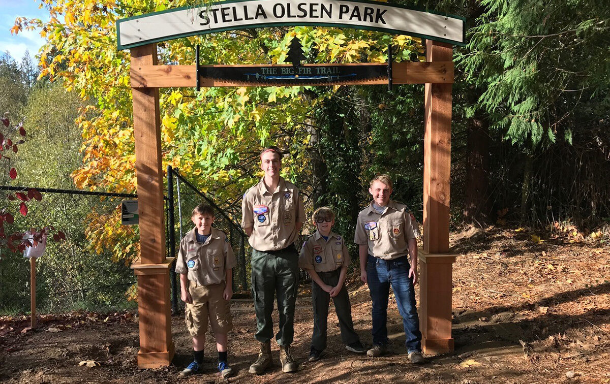 Four people in Scouting uniforms stand under the entrance sign of Stella Olsen Park, surrounded by trees with autumn foliage.
