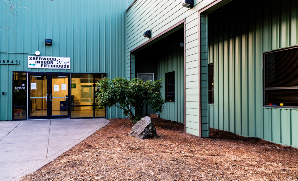Entrance to a green metal building labeled "Sherwood Indoor Fieldhouse," with double glass doors and a small landscaped area in front.