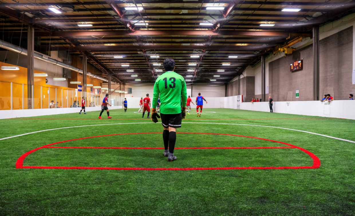 An indoor soccer goalkeeper in a green jersey stands near the goal as players in various colored uniforms compete on the field.